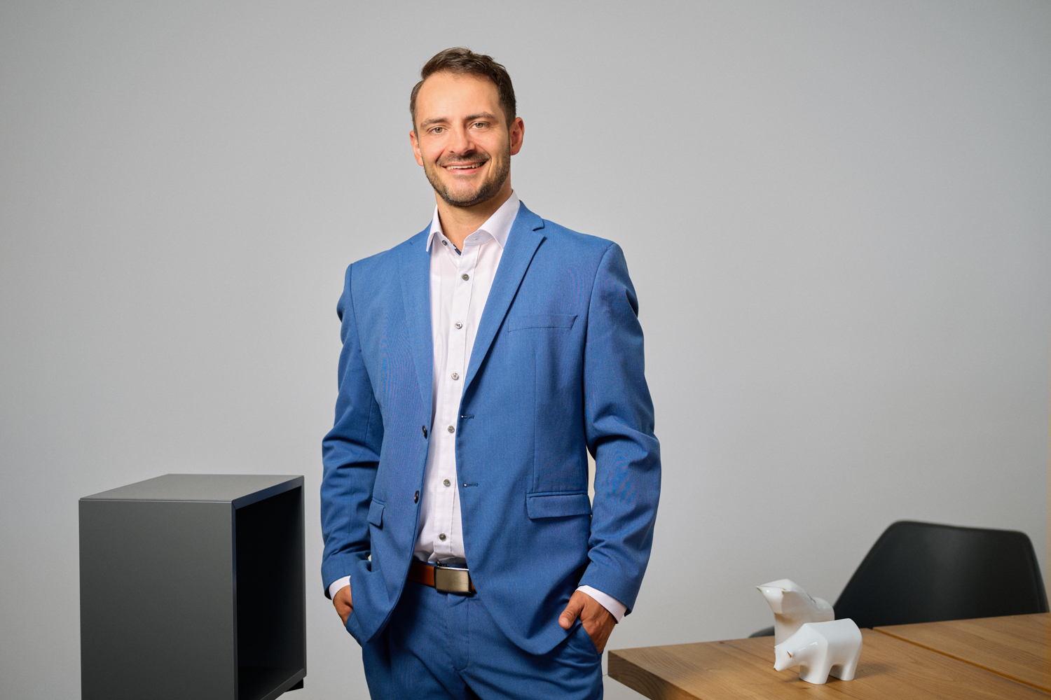 Portrait von Alexander Stengel - Vorstand der Fortezza Finanz AG in Nürnberg: Portrait von Wolfgang Dirsch - Vorstand der Fortezza Finanz AG in Nürnberg: ein junger Mann mit dunklem Haar und Bart in hellblauem Anzug mit weißem Hemd steht lächelnd in einem modernen Büro. Seine Hände sind in den Hosentaschen, im Hintergrund ein Tisch mit weißen Stier- und Bärfiguren.