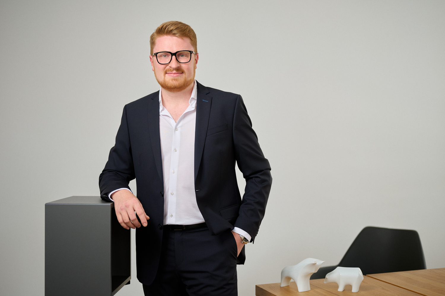 Portrait von Benno Roggenhofer: ein junger Mann mit kurzem roten Haar und Bart, gekleidet in einen dunkelblauen Anzug mit weißem Hemd, lehnt entspannt an einem Möbelstück in einem modernen Büro. Im Hintergrund stehen weiße Stier- und Bärfiguren auf einem Holztisch.